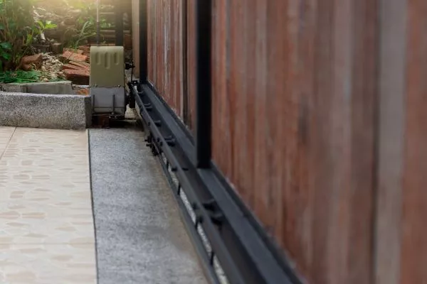 A close-up view of an automated sliding gate with a wooden finish, mounted on a metal rail along an Austin stone-paved driveway. There's a small motor at the end, surrounded by some greenery and a concrete platform in the background.