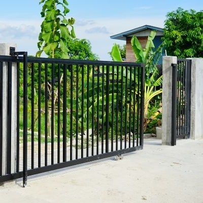 A black metal gate stands between two concrete pillars, bordered by lush green plants and trees. In the background, a small wooden structure is visible, set against a blue sky with some clouds. The ground around the gate is paved with concrete, representing the community's pride in supporting local craftsmanship.