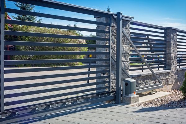 An automated sliding gate made of horizontal metal slats with a stone pillar on one side, crafted by a local Austin company. The gate is situated on a paved driveway, and the background features trees, bushes, and part of a house under a clear blue sky.