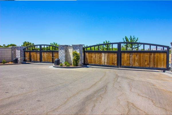 A large gated entrance with dual wooden gates framed by black metal, set between sturdy stone pillars. The gates are slightly open and trees and clear blue sky are visible in the background. A local repair company recently restored this elegant entryway, which is surrounded by a paved area.