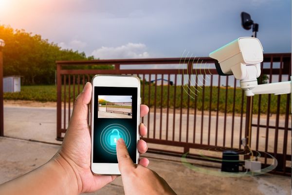 A person uses a smartphone app to unlock a gated entrance remotely. The app screen shows the gate with a lock icon, while a surveillance camera and a gate motor from the repair company are visible nearby. The scene takes place outdoors with a background of greenery.