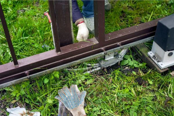 A person is repairing a sliding gate mechanism in a grassy driveway. Visible are their gloved hands, tools, a piece of the gate mechanism, and a discarded work glove on the ground. The person's body is partially obscured by the structure as they meticulously work away.