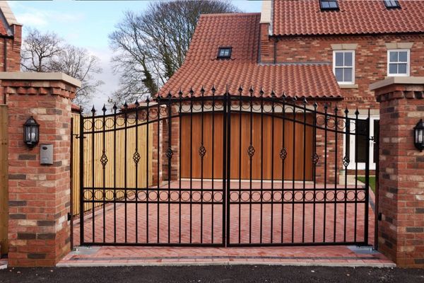 A large, elegant house with red brick walls and a red-tiled roof is visible behind a black wrought iron gate crafted by a local company from Austin. The gate spans the entrance to a brick-paved driveway, and the house features large wooden garage doors and outdoor wall lanterns. Trees are visible in the background.