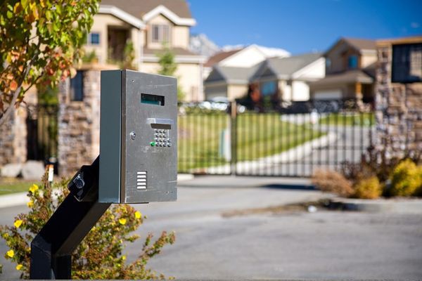 A close-up of a gated community's access control keypad under a clear blue sky, with suburban houses and a closed gate in the background. The keypad is mounted on a black post and surrounded by trees and shrubs, providing an inviting welcome that consistently impresses residents and visitors alike.