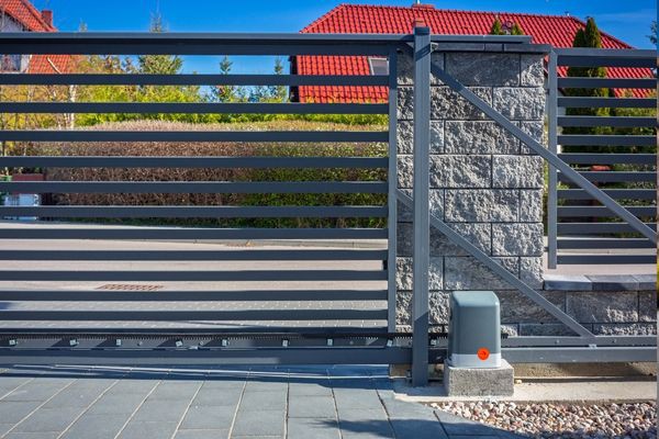 A modern, horizontal slat metal gate with a motorized sliding mechanism is shown, mounted between stone pillars. Crafted by a local company, the sleek design complements the house with a red-tiled roof and well-maintained yard visible behind it. The sky is clear and blue.