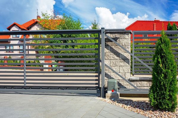 A modern metal fence with horizontal slats sits in front of a house with red roofs and lush greenery in Austin. The fence includes a sliding gate and a small section of stone wall. There are shrubs and a paved driveway in the foreground, set against a cloudy blue sky, ready for any local repair needs.