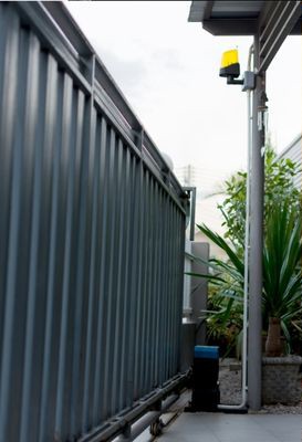 A metal fence with vertical bars is partially open next to a paved walkway leading to a driveway. A small motor is attached to the base of the fence, possibly awaiting repair. On a nearby pole, a yellow flashing light is mounted. In the background, there's a potted plant and part of a building's exterior.