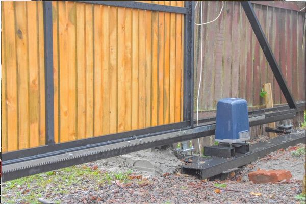 An automatic sliding gate in a residential area. The gate features horizontal wooden slats framed by black metal, seamlessly blending with the driveway. The mechanism for the gate is visible, including a blue motor unit and a gear drive on a metal track, with a brown fence in the background.