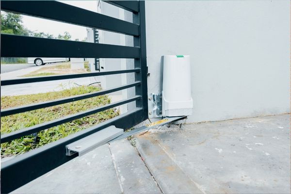 A black metal gate with an automatic sliding mechanism is attached to a concrete wall. Near the ground, a white control box manages the operation. In the background, a grassy area and part of a road with a white car are visible, reflecting local Austin charm and reliability in repair services.