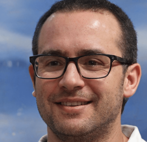 A person with short dark hair, glasses, and facial stubble smiles against a blue sky background. They are wearing a white shirt, standing near a freshly repaired driveway.