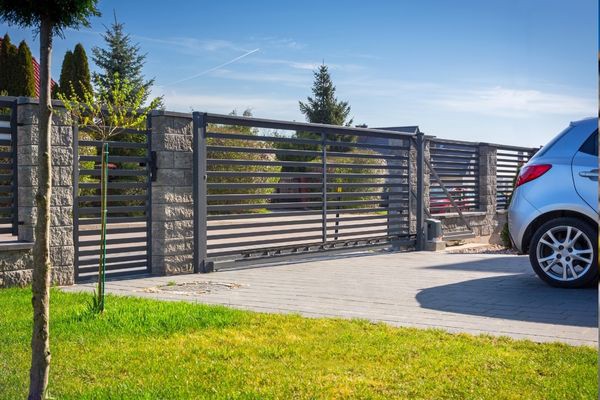 A metallic sliding gate and stone pillars mark the entrance to a property, with a silver car partially visible on the right. The driveway leads to a grassy area with a young tree in the foreground, and a clear blue sky with a plane streak in the background.