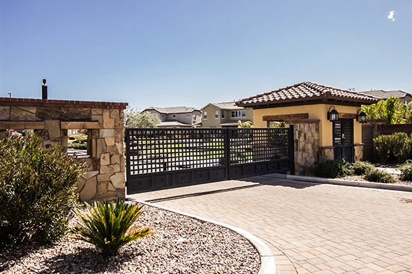 A gated entrance to a residential community with a black metal gate, stone walls, and a tiled roof guardhouse marks the start of the scenic driveway. The area is landscaped with various plants and trees, and several houses are visible in the background under a clear blue sky.