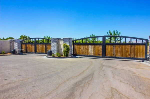 A gated entrance with large dual sliding wooden gates bordered by black metal frames, set into a stone wall structure. The surrounding area is paved, with small landscaped spots featuring a few plants. The sky is clear and blue, perfect for a local repair team in Austin to work efficiently.