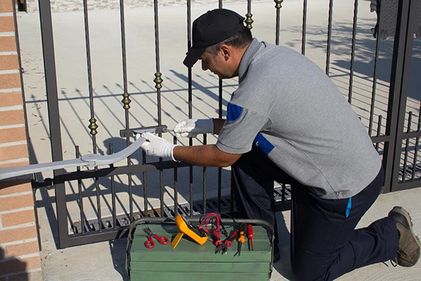 A local man in a gray uniform and black cap kneels next to an iron gate, working with tools to repair it. He wears white gloves and holds a tool in his hands. A green toolbox with various tools is open on the ground beside him. The setting appears to be outdoors, perhaps in Austin.