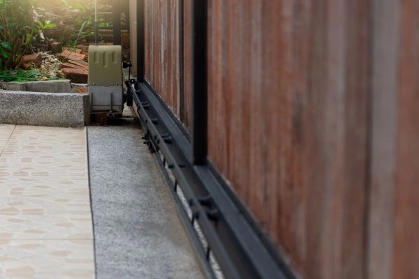 A close-up view of an automated sliding gate with a wooden finish, mounted on a metal rail along an Austin stone-paved driveway. There's a small motor at the end, surrounded by some greenery and a concrete platform in the background.