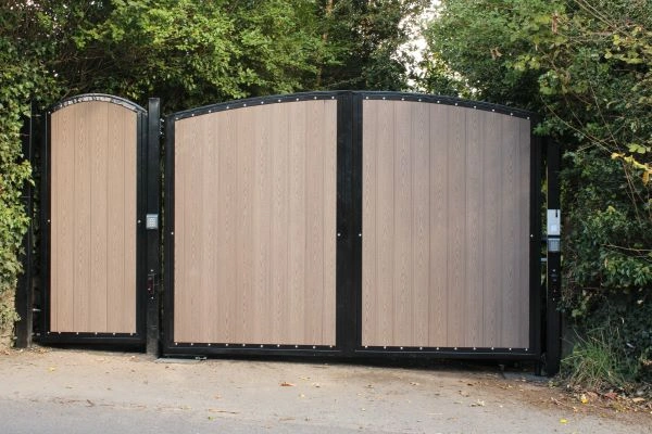 A large, double panel, wooden gate with a smaller pedestrian gate attached on the left side, framed with a dark material. Surrounded by greenery including bushes and trees, this charming entrance appears to be at a private property in Austin. Perfect for any company specializing in bespoke repair services.