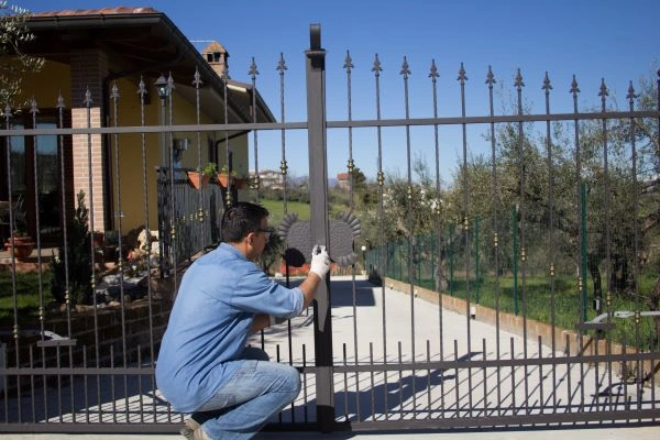 A man in a blue shirt and gloves is kneeling and working on a metal gate with a screwdriver outside a house. The house, featuring a brick exterior and tiled roof, has potted plants hanging nearby. The driveway beyond the gate extends toward the open space of the company's outdoor area.