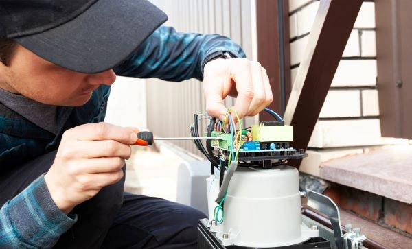 A person wearing a cap and plaid shirt is using a screwdriver to work on the wiring of an electrical device outdoors. The device, mounted on a metallic base, sits near the driveway. Various colored wires are visible against the backdrop of a brick wall and wooden fence.