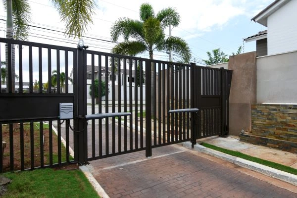 A black metal gate with vertical bars, crafted by a local company in Austin, surrounds a residential compound. Featuring an automated sliding mechanism, the gate opens onto a brick-paved driveway, with palm trees and modern houses visible in the background.