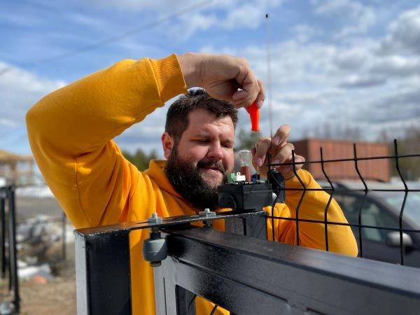 A man in a yellow hoodie is using a red screwdriver to repair a metal component on a black wire gate. The background shows a partly cloudy sky, some trees, and various outdoor equipment.