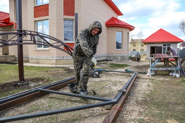 A person in camouflage clothing works on constructing a metal frame outside a brick house with red roofs. Positioned on the gravelly ground by the driveway, they are surrounded by various building materials and tools. A gate and another building are visible in the background.