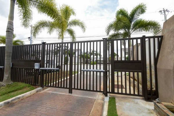 A brown metal gate with vertical slats stands open, flanked by concrete walls and tall palm trees. Behind the gate, a paved driveway leads into a residential or commercial area in Austin. The sky is partly cloudy, and power lines are visible in the background, hinting at potential repair work needed by the company.