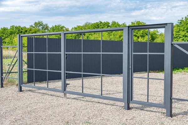 A sleek, modern metal gate with a symmetrical grid design stands closed on a gravel driveway. The gate is framed by a tall, solid dark-colored fence, blending with the lush green trees and blue sky in the background.