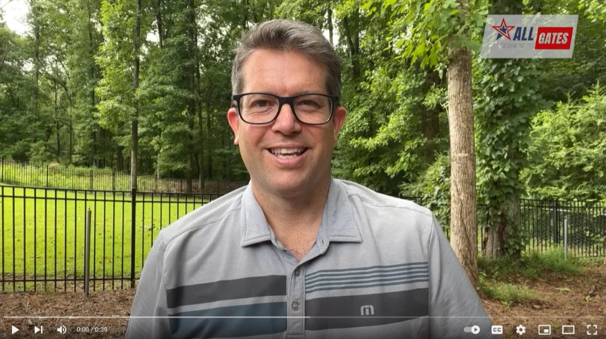 A man wearing glasses and a light blue polo shirt with a gray stripe stands smiling in a wooded outdoor area. The background features trees, a metal fence, and a grassy area. In the top right corner, a logo reads "All Gates," indicating the local company's presence in Austin.